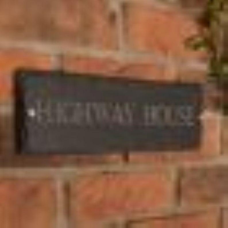 May include: A rectangular black sign with the words "HIGHWAY HOUSE" in white lettering is mounted on a brick wall. The sign is slightly weathered, suggesting outdoor use. The brick wall has a reddish-brown color.