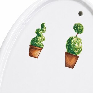 May include: White oval serving platter with a decorative garden theme. Features two potted topiary plants in brown pots with green foliage. The platter has a small hole near the top edge.