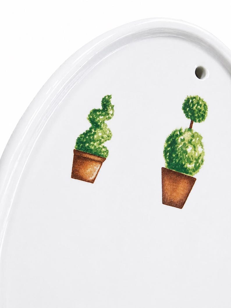 May include: White oval serving platter with a decorative garden theme. Features two potted topiary plants in brown pots with green foliage. The platter has a small hole near the top edge.