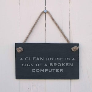 Pode incluir: Uma placa retangular de ardósia preta com o texto "A CLEAN HOUSE IS A SIGN OF A BROKEN COMPUTER" em letras cinza claro. A placa é suspensa por uma corda natural e está pendurada em um fundo de madeira branca.
