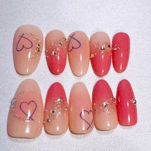 May include: A set of ten oval-shaped press-on nails featuring a gradient transition from light peach to vibrant pink. The nails are decorated with hand-painted purple heart outlines, scattered gold glitter accents, and small clear rhinestones.