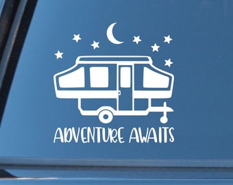 May include: White vinyl decal of a camper trailer with a crescent moon and stars above it. The text "Adventure Awaits" is below the camper. The decal is on the back window of a black car.