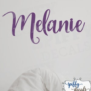 May include: Purple vinyl wall decal with the name "Melanie" in a cursive font. The decal is on a white wall. The text "spiffy decals" is in a black frame with a white background.