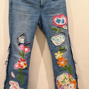 May include: Light blue denim jeans with colorful, whimsical patches. Patches include flowers, a dog, and the text "BOW WOW" and "RUFF". The jeans have a frayed edge detail along the outer seams.