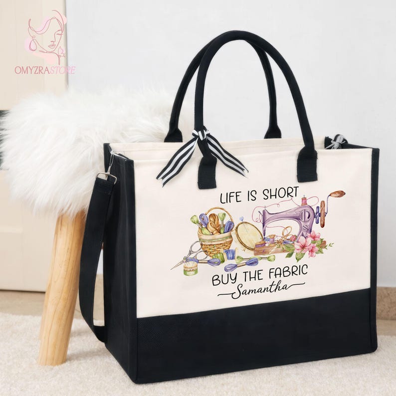 May include: A black and white tote bag with black handles and a shoulder strap. The bag features a sewing-themed design with text that reads "LIFE IS SHORT BUY THE FABRIC Samantha". The design includes a sewing machine, flowers, and sewing supplies.