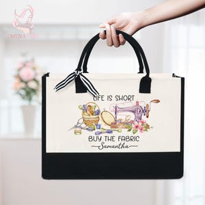 May include: A black and cream tote bag with black handles and a decorative bow. The bag features a sewing-themed design with text that reads "LIFE IS SHORT BUY THE FABRIC Samantha".