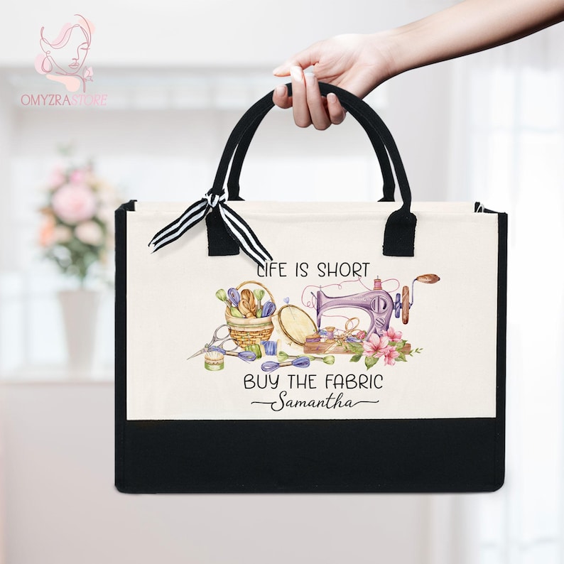 May include: A black and cream tote bag with black handles and a decorative bow. The bag features a sewing-themed design with text that reads "LIFE IS SHORT BUY THE FABRIC Samantha".
