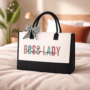 Peut inclure: Un sac fourre-tout blanc avec des poignées noires et une base noire. Le sac présente le texte "BOSS LADY Katrina" en lettres colorées et un nœud rayé noir et blanc.
