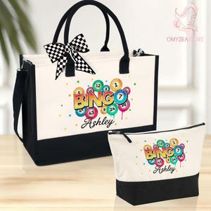 May include: A tote bag and matching cosmetic bag set. The bags are white with black accents and handles. The tote bag has a black and white chequered bow. Both bags feature a colourful "Bingo Ashley" design with bingo balls.