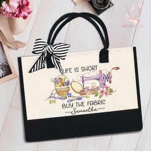 May include: A canvas tote bag with black handles and a black bottom panel. The bag features a watercolor design of a sewing machine, flowers, and text that reads "Life is Short, Buy the Fabric Samantha". A black and white striped bow is attached.