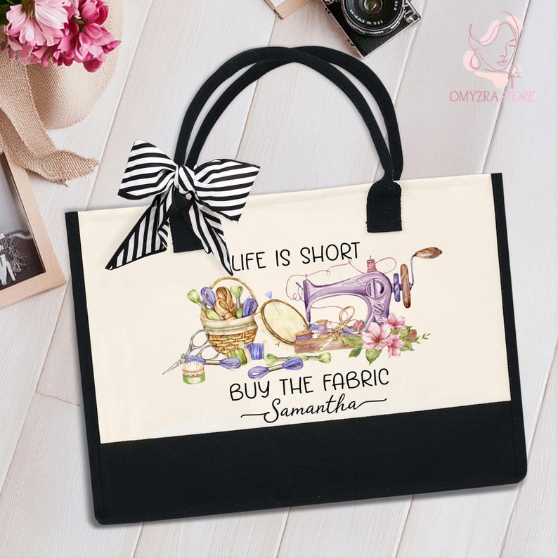 May include: A canvas tote bag with black handles and a black bottom panel. The bag features a watercolor design of a sewing machine, flowers, and text that reads "Life is Short, Buy the Fabric Samantha". A black and white striped bow is attached.