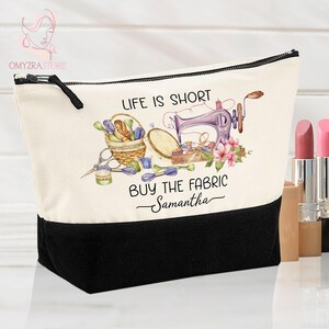 May include: A canvas cosmetic bag with a black base and a cream-colored top. The bag features a sewing-themed design with a sewing machine, basket of supplies, and the text "Life is Short, Buy the Fabric, Samantha."