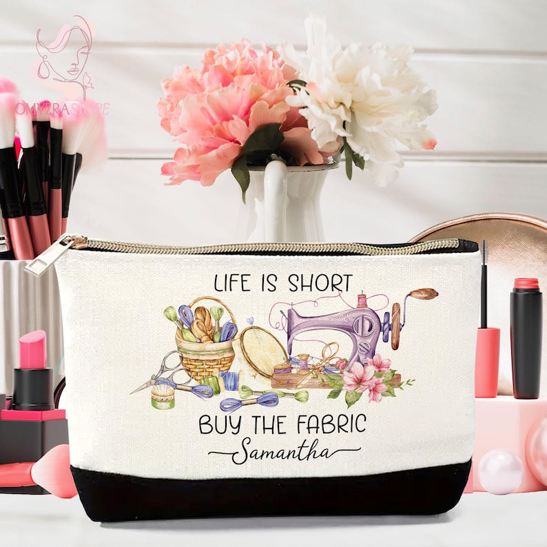 May include: A white cosmetic bag with a black bottom and a gold zipper. The bag features a sewing-themed design with a sewing machine, basket of thread, and the text "LIFE IS SHORT BUY THE FABRIC Samantha".