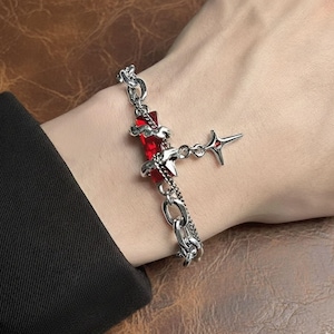 Può includere: Un braccialetto argentato con maglie a catena e dettagli di pietre preziose rosse. Il braccialetto presenta una pietra rossa rettangolare centrale e un ciondolo a forma di stella con un centro rosso. Il braccialetto è indossato al polso.