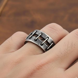 Op de afbeelding: Een zilverkleurige ring met een uniek ontwerp. De ring heeft een reeks parallelle staven die zijn verbonden door verticale lijnen, waardoor een gestikt of geregen uiterlijk ontstaat. De ring wordt aan een vinger gedragen.