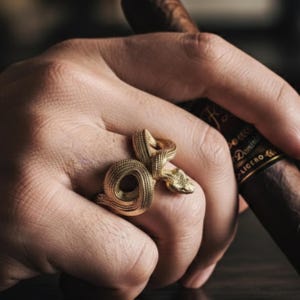 May include: A gold-colored snake ring with detailed scales and a coiled body. The ring is worn on a hand, with a cigar in the background. The ring's design features a snake head and a textured surface.