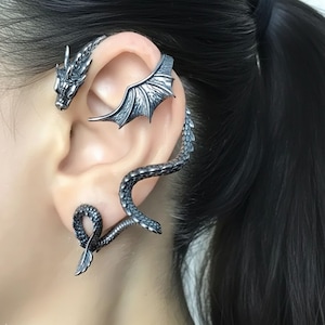 May include: A silver-toned dragon ear cuff. The cuff wraps around the ear, featuring a dragon head, wing, and a serpentine body that curves down. The dragon's scales and details are clearly visible.