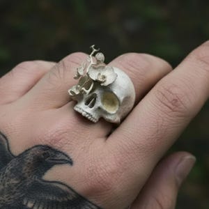 May include: A detailed, off-white skull ring with floral accents. The ring features a skull design with eye sockets and teeth, and delicate flowers on top. The ring is a unique piece of jewelry.