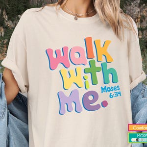 Könnte beinhalten: Cremefarbenes T-Shirt mit dem Text "Walk With Me" in bunten Buchstaben. Das Design beinhaltet ein Kreuz und den Verweis "Moses 6:34". Teil der Comfort Colors Kollektion.