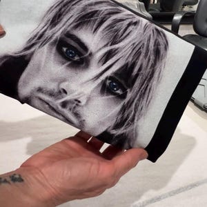 May include: Black and white portrait print on a rectangular fabric. The image features a close-up of a person's face with blue eyes and light-colored hair. The fabric has a black border.
