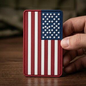 May include: Rectangular patch featuring the American flag design. The patch has a red border, with red and white stripes on the left and a blue field with white stars on the right. The patch is held by a hand.