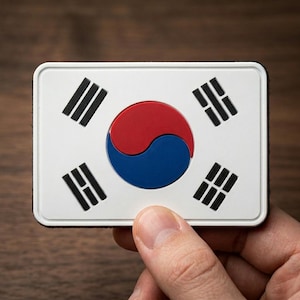 May include: A rectangular patch featuring the South Korean flag. The flag design includes a white background, a central red and blue Taegeuk symbol, and four black trigrams in each corner. The patch is held by a hand.