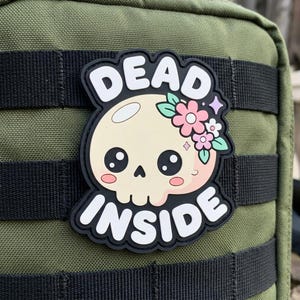 May include: A black and white patch with the words "DEAD INSIDE" surrounding a cartoon skull with pink cheeks, large eyes, and a floral crown. The patch is affixed to a green fabric background.