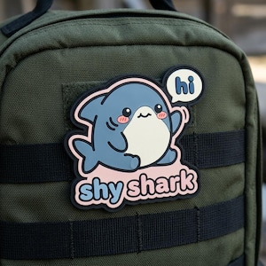 May include: A patch featuring a cartoon shark with a light blue body, white belly, and pink cheeks. The patch has the text "shy shark" and a speech bubble that says "hi". The patch is attached to a green backpack.