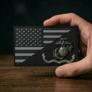 May include: A rectangular black and grey patch featuring a stylized American flag design divided diagonally. The lower right section displays the United States Marine Corps emblem, which includes an eagle, globe, anchor, and a banner reading UNITED STATES MARINE CORPS.