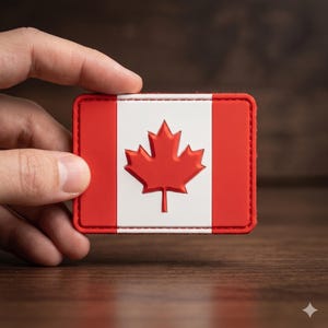 May include: A rectangular patch featuring the Canadian flag. The patch has a red border and a white center with a red maple leaf. The background is a blurred wooden surface.