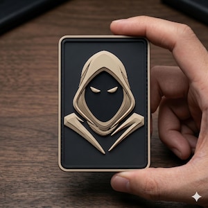 May include: A rectangular patch featuring a stylized hooded figure with a black face and two white eyes. The hood and surrounding details are tan, set against a black background. The patch has a tan border.
