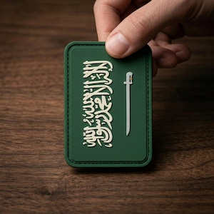 May include: A green rectangular patch featuring the flag of Saudi Arabia. The flag design includes white Arabic script and a white sword. The patch is held against a dark wood grain surface.