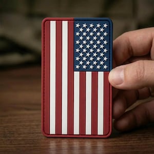 May include: A rectangular patch featuring a raised, molded design of the United States flag. The design includes vertical red and white stripes on the left and a blue field with white stars on the right, all rendered in a durable, textured material.