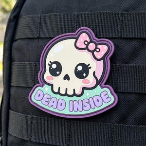May include: A cute skull patch with a pink bow and the words "DEAD INSIDE" in a bubble font. The skull has large, expressive eyes and pink cheeks. The patch is outlined in purple and set against a black background.