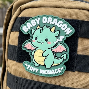 May include: A green and yellow "Baby Dragon" patch with the text "Tiny Menace". The patch features a cartoon dragon with pink wings and a wisp of smoke. The patch is attached to a black velcro square on a tan backpack.