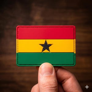 May include: A rectangular patch featuring the flag of Ghana. The patch has a red, yellow, and green horizontal stripe design with a black star in the center. The patch is held by a hand.