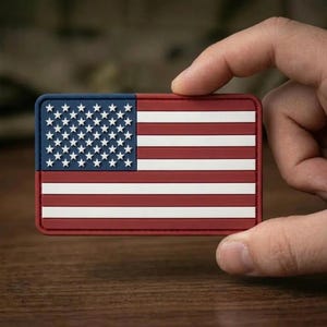 May include: A rectangular patch featuring the United States flag design with a dark blue canton containing raised white stars and alternating horizontal red and white stripes. The patch has a smooth, molded texture with clean, defined edges.