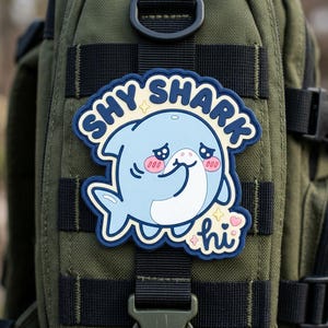 May include: A blue and white patch featuring a cartoon shark with the text "SHY SHARK" and "hi". The shark has blushing cheeks and is smiling. The patch is attached to a green backpack.