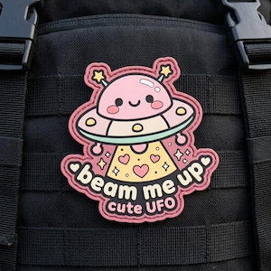 May include: A pink and cream-colored patch featuring a cute UFO design with the text "beam me up cute UFO." The patch has a smiling face and star accents. It is attached to a black tactical vest.