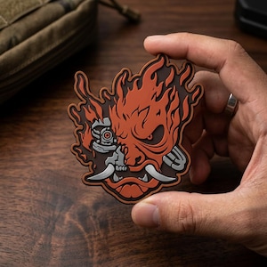May include: A rubberized morale patch featuring a stylized oni mask design in orange and dark brown. The graphic depicts a half-mechanical skull with a glowing red eye and sharp silver fangs, surrounded by flame-like shapes on a dark background.