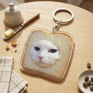 May include: A clear acrylic keychain featuring a white cat's face framed by a slice of bread. The cat has blue eyes and a pink nose. The keychain is attached to a silver metal ring.