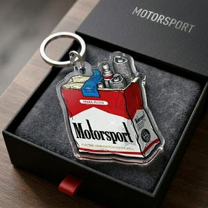 Puede incluir: Un llavero Motorsport con un dise&ntilde;o que se asemeja a un paquete de cigarrillos, con buj&iacute;as. El llavero es rojo y blanco con la palabra "Motorsport" y el texto "Electric your ignition system well". Se presenta en una caja de regalo negra.