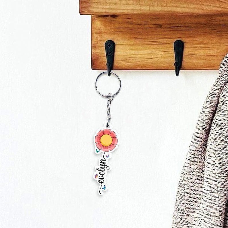 Puede incluir: Un llavero floral con un dise&ntilde;o de flor roja y amarilla. El llavero tiene el nombre "evelyn" escrito en una fuente de escritura. El llavero est&aacute; colgado de un gancho de metal en un estante de madera.