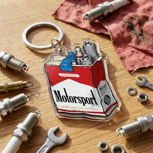 Puede incluir: Un llavero acr&iacute;lico transparente con un dise&ntilde;o rojo y blanco de "Motorsport", que se asemeja a un paquete de cigarrillos, que contiene buj&iacute;as. La imagen incluye buj&iacute;as, llaves y otras herramientas automotrices.