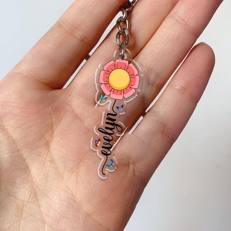 Puede incluir: Un llavero de acr&iacute;lico transparente con un dise&ntilde;o de flor rosa y el nombre "evelyn" en escritura cursiva. La flor tiene un centro amarillo y est&aacute; adornada con peque&ntilde;as mariposas azules. El llavero est&aacute; sujeto a una cadena plateada.
