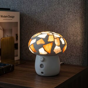 Puede incluir: Una lámpara de mesa con forma de seta, base blanca y pantalla estilo vidriera con piezas geométricas translúcidas en color ámbar y blanco. La lámpara emite una luz cálida desde el interior de la pantalla, y la base incluye un interruptor circular y dos pequeñas hendiduras.
