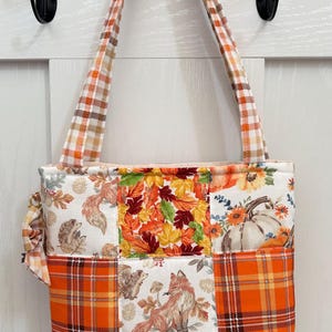 May include: A patchwork tote bag featuring a seasonal autumn theme with orange, yellow, and brown plaid fabric squares interspersed with panels depicting foxes, hedgehogs, pumpkins, and fall leaves. The bag has two matching plaid shoulder straps.