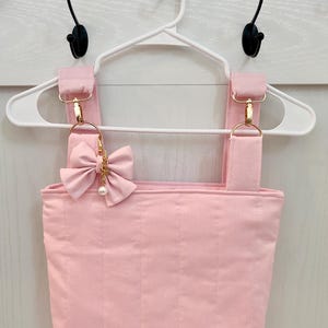 May include: A light pink quilted top featuring wide shoulder straps attached with gold-toned metal lobster clasps and rings. A decorative pink fabric bow with a small dangling gold chain and a white faux pearl charm is attached to the right strap.