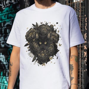 Puede incluir: Camiseta blanca con un diseño artístico de un gato negro con ojos amarillos, rodeado de flores y mariposas negras con detalles dorados. El diseño es detallado. Cuello redondo.
