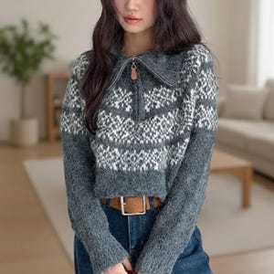 Peut inclure: Un pull gris et blanc à motifs avec un col zippé et une tirette en cuir marron. Le pull présente un motif géométrique et est associé à une ceinture marron et un jean bleu. Le pull est de style court.